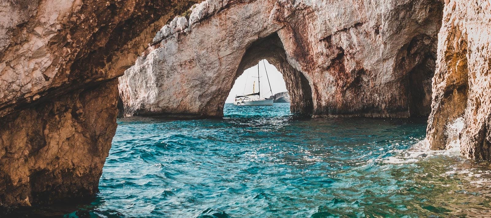 A sailboat passing through a rock arch on blue sea